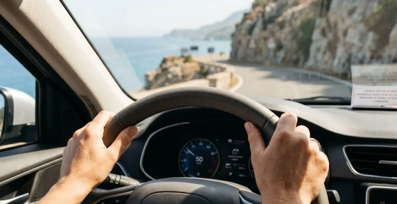 Dettaglio ravvicinato di mani sul volante di un'auto moderna, sullo sfondo sfocato si intravede una strada costiera sarda con paesaggio mediterraneo